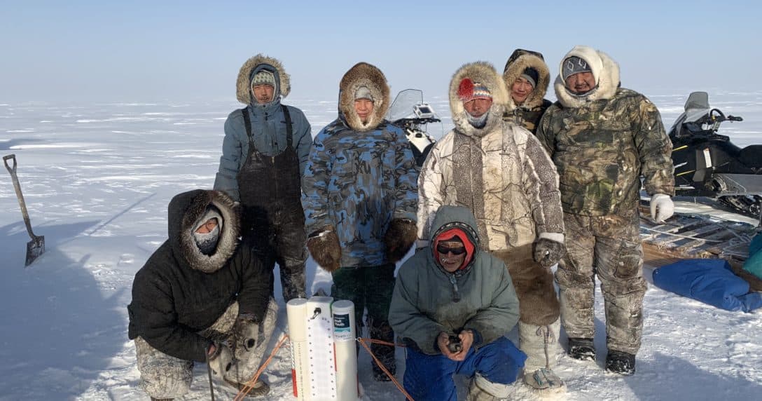 Image description: Seven community members and SmartICE operators in Igloolik, Nunavut are deploying a SmartBUOY device in the sea ice in March 2020. Data from the SmartBUOY is transmitted daily via satellite and made available to the community on SIKU.org.
