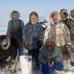 Image description: Seven community members and SmartICE operators in Igloolik, Nunavut are deploying a SmartBUOY device in the sea ice in March 2020. Data from the SmartBUOY is transmitted daily via satellite and made available to the community on SIKU.org.
