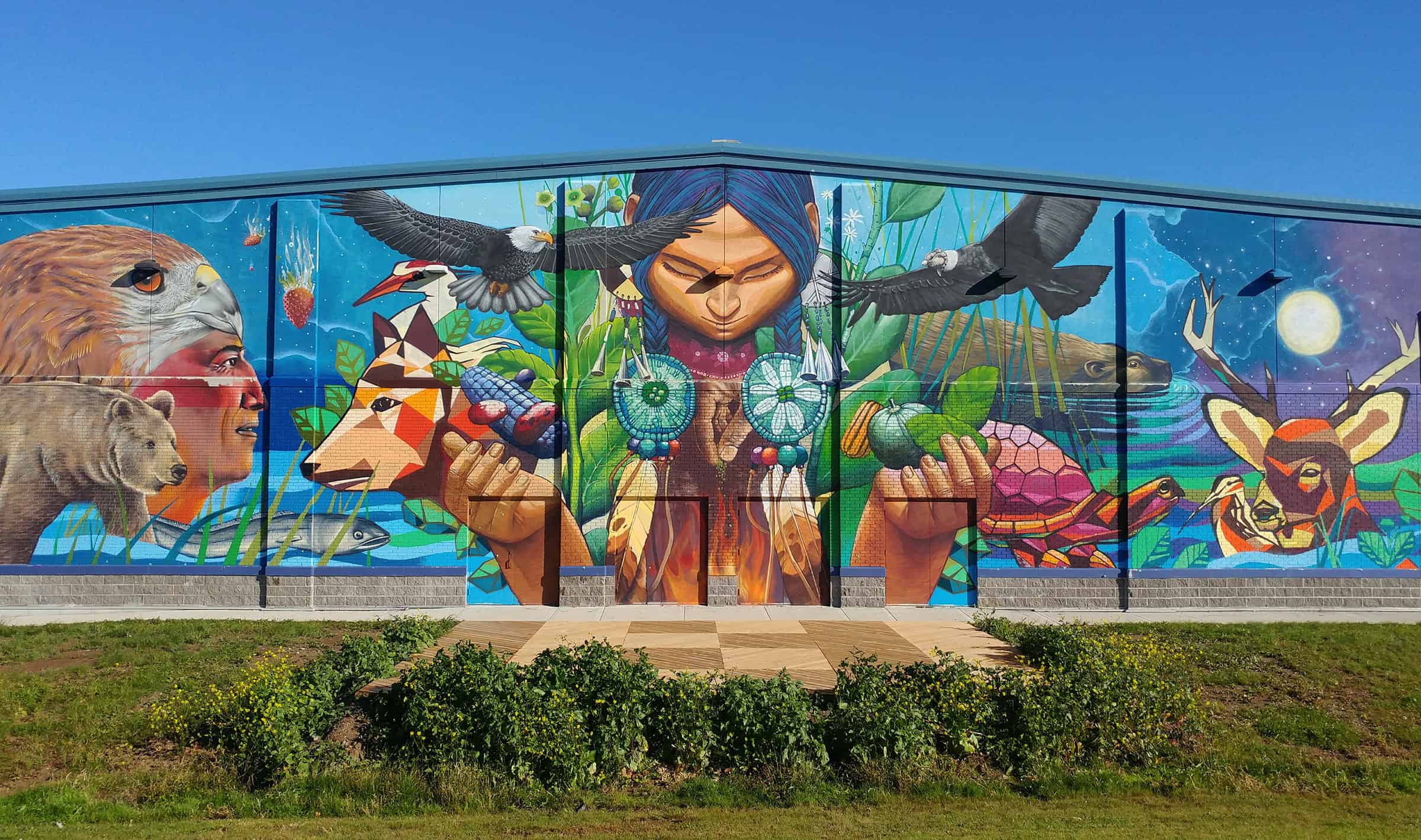 A colourful mural that takes up the entire side of a large building. A person at the centre with animals surrounding them.