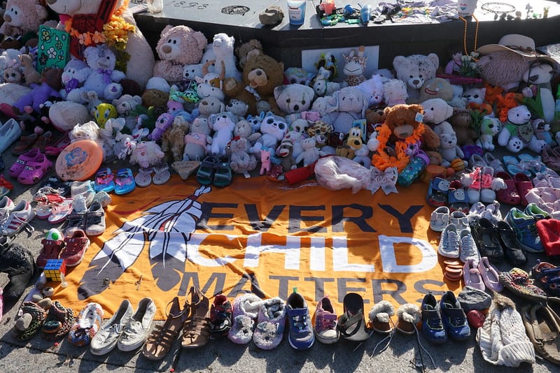 Memorial with flag placed on the ground that reads "Every Child Matters" with children's shoes and stuffed animals surrounding it.