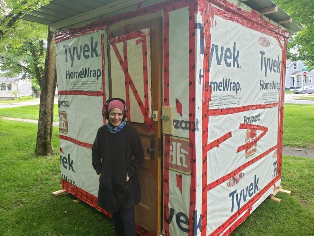 Person smiling in front of a small, temporary home.