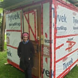 Person smiling in front of a small, temporary home.
