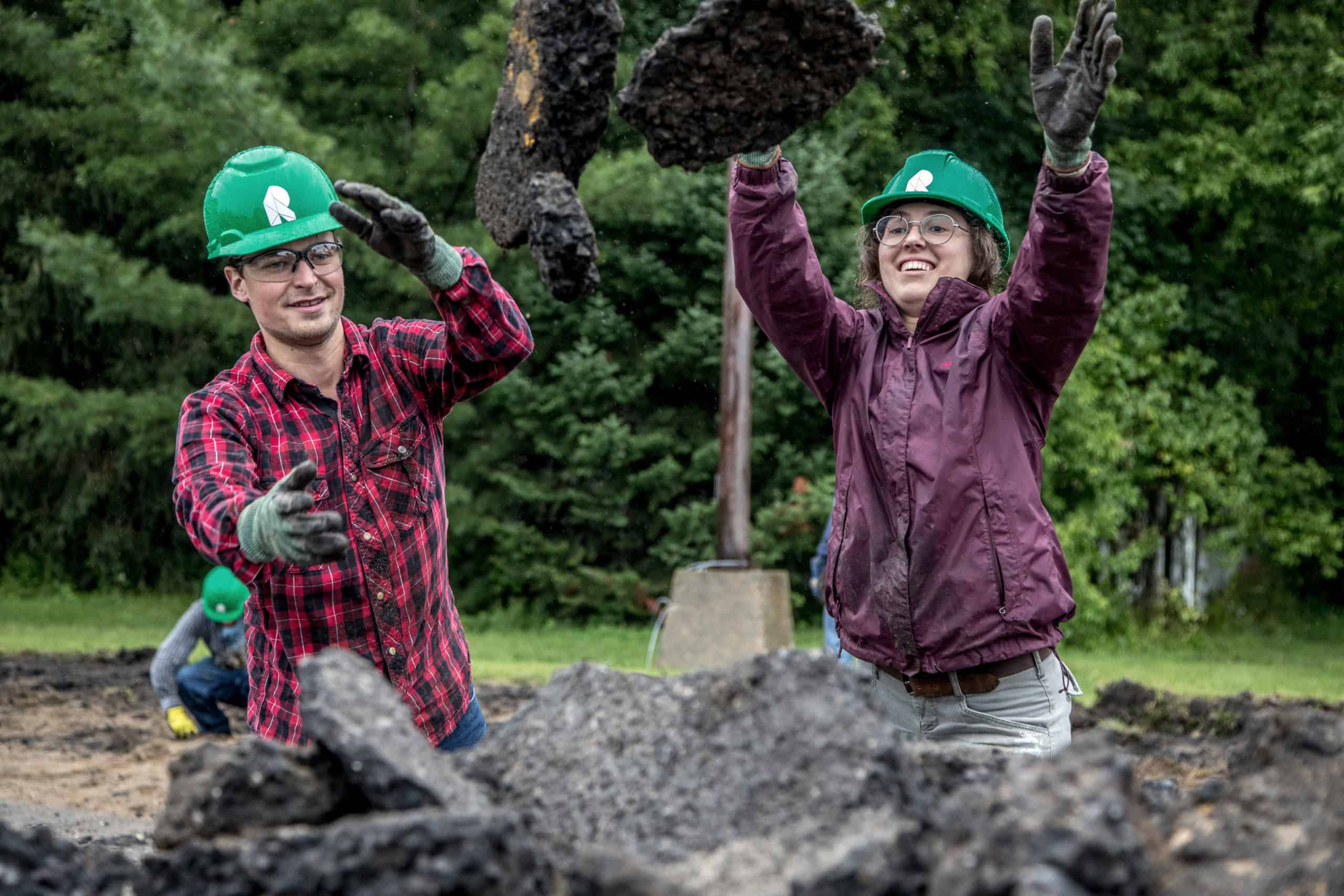 Two volunteers with hard hats throw a piece of asphalt into a bin.
