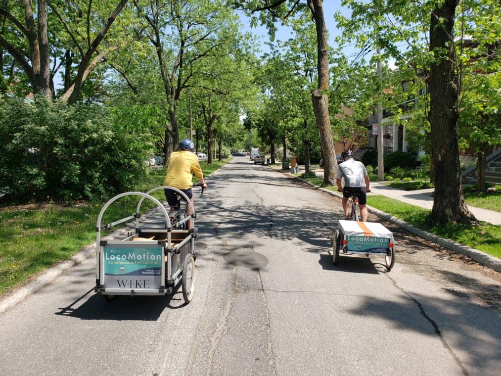 Two people cycling on a street pulling trailers that have a sign that reads, "LocoMotion."
