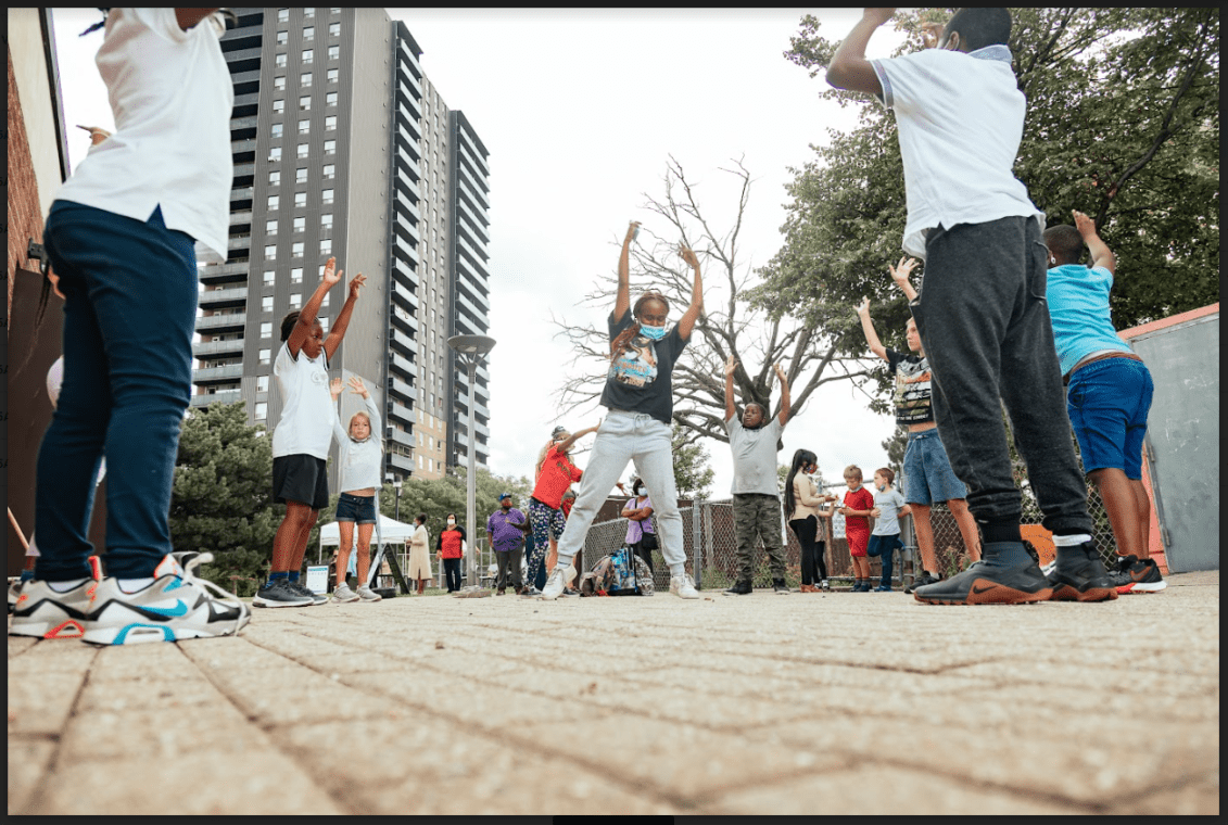 A photo of kids dancing outside with their arms in the air. A high rise building is in the distance.