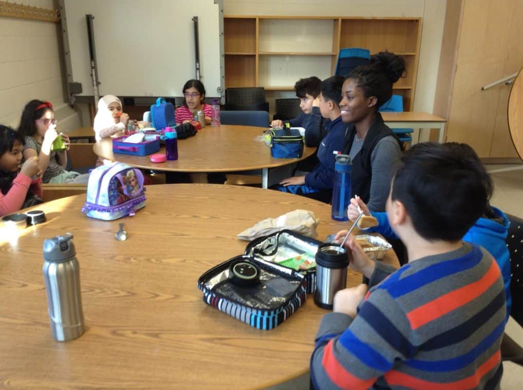 Kids eating lunch out of lunch boxes and water bottles at tables with an adult sitting with them.