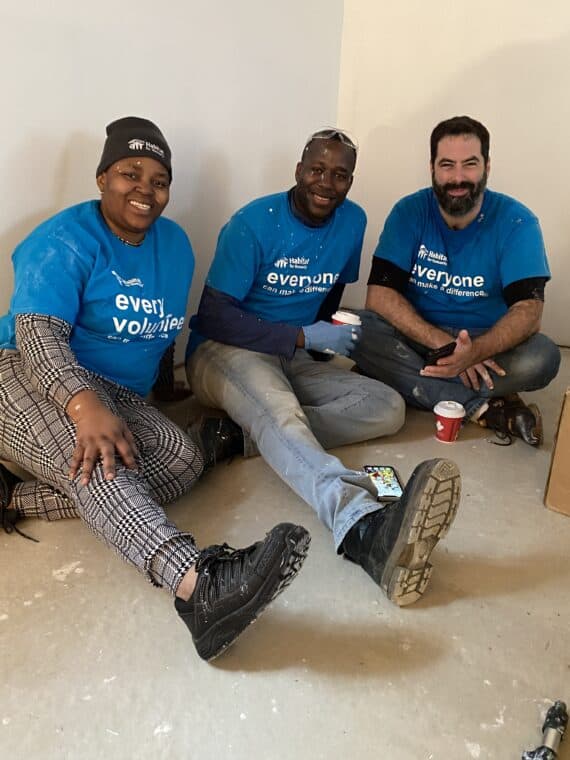 Three people sitting at a build site wearing matching t-shirts that says "everyone volunteers"