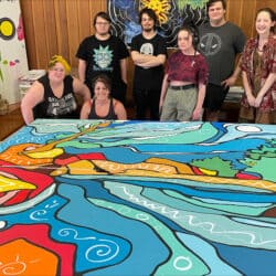 Seven young people smiling behind a large colourful painting.