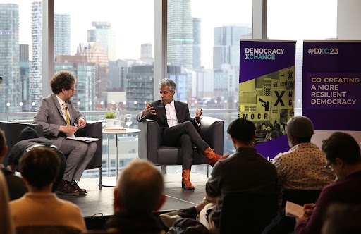 Anand Giridharadas speaking on stage. A sign next to him says "Democracy Xchange"