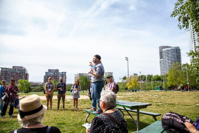 A person standing on a picnic table is speaking to a group in a green space outside.