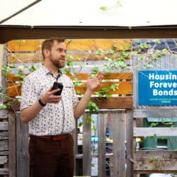 Person speaking under a garden tent. There is a sign in the background that says "Housing Forever Bonds"