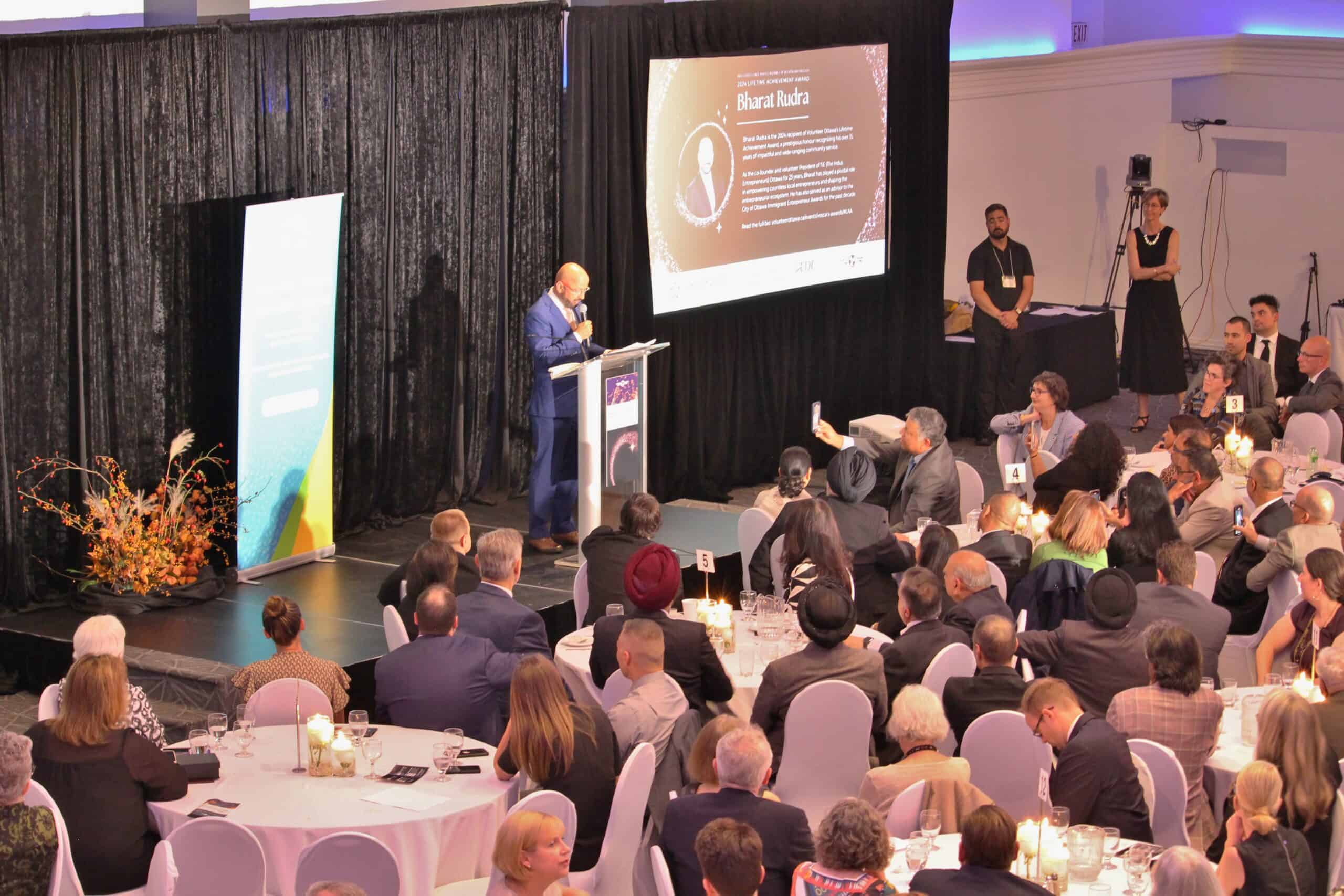 Someone at a podium with filled tables of people around them. There is a slide projected with the name "Bharat Rudra"