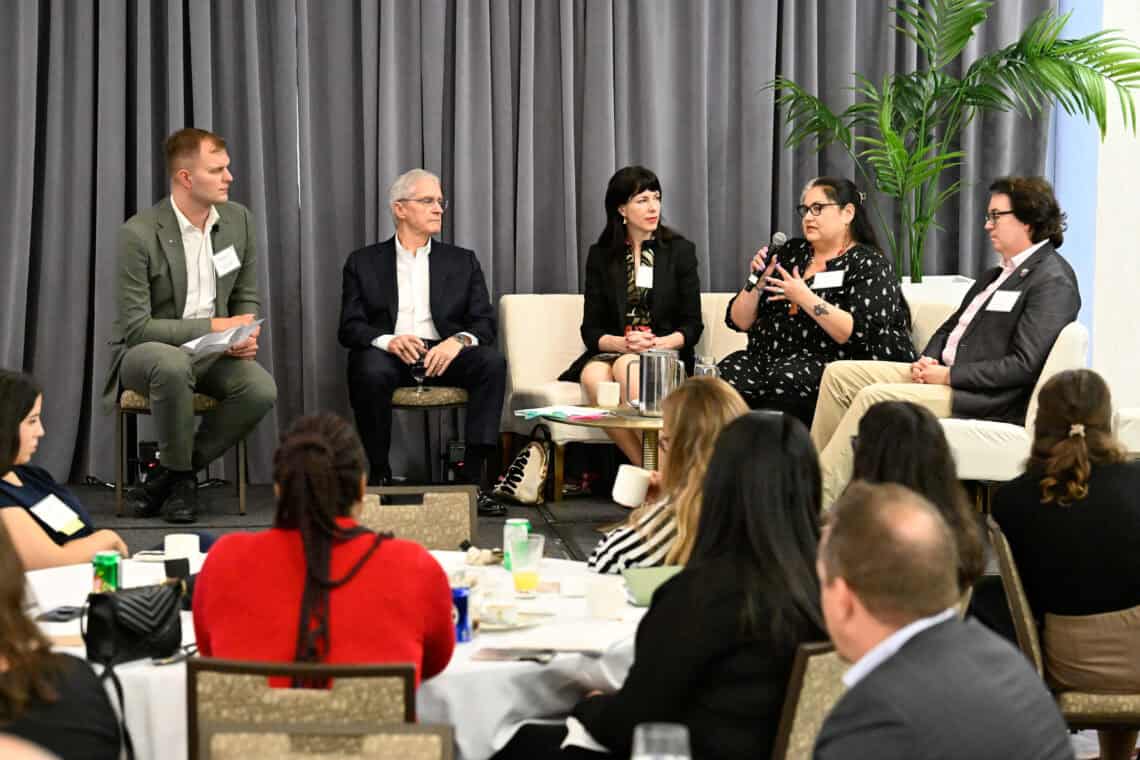 Five people seated around a coffee table on a stage, watching one person speak with a microphone.