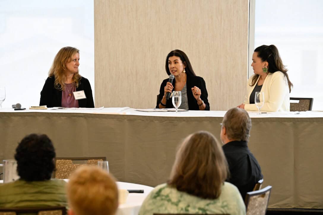 Three people sitting as panelists at a table. The person in the middle is speaking into a microphone.