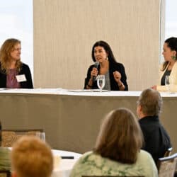 Three people sitting as panelists at a table. The person in the middle is speaking into a microphone.