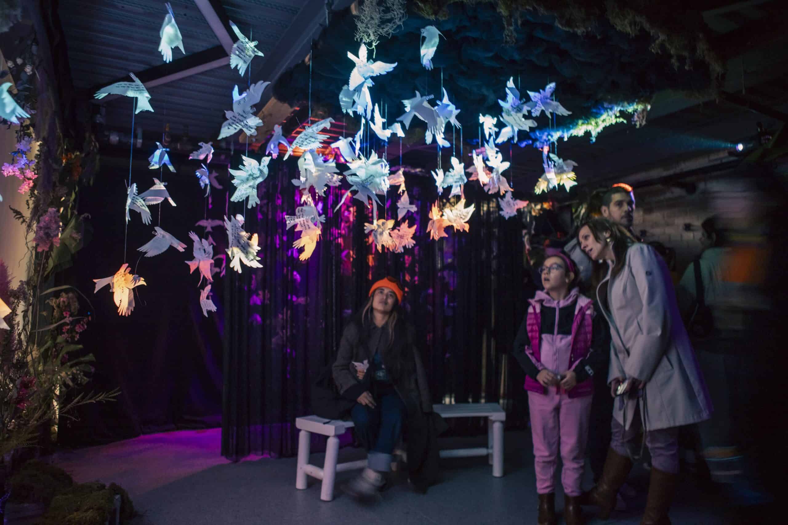 An intergenerational group of people stand inside Space for Grief watching a film projected onto paper birds that act as both canvas and sculpture, casting a shadow of a murmuration onto the screen behind it.