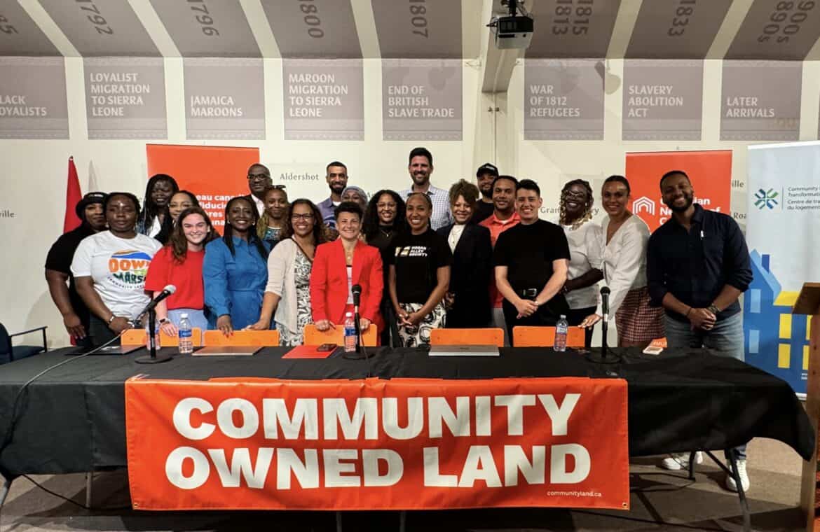 A group of 22 people stand behind a table that says "Community Owned Land"