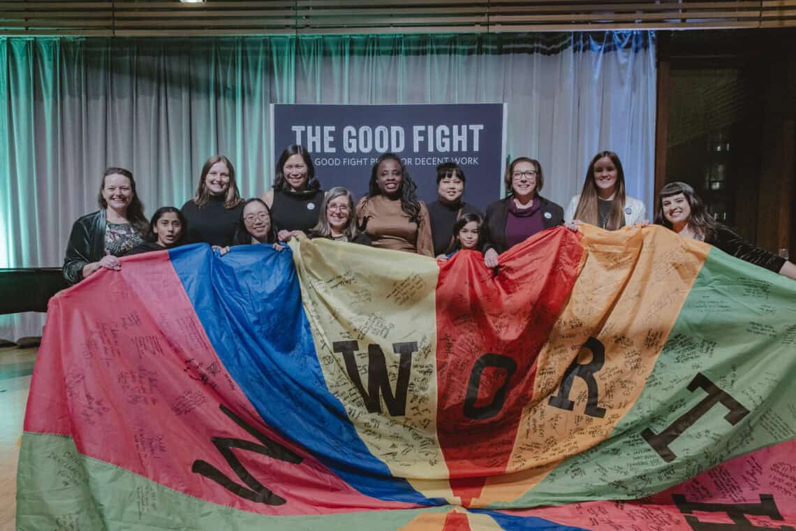 12 people smiling and posing in front of a sign that reads "The Good Fight Prize for Decent Work" and hold a colourful parachute that has letters and signatures or small writing on it.