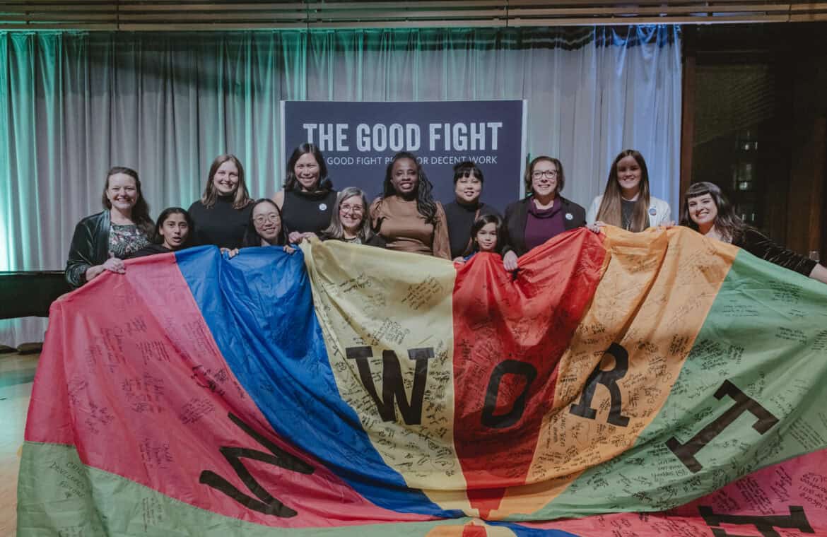 12 people smiling and posing in front of a sign that reads "The Good Fight Prize for Decent Work" and hold a colourful parachute that has letters and signatures or small writing on it.