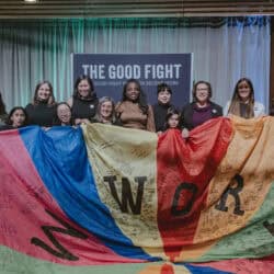 12 people smiling and posing in front of a sign that reads "The Good Fight Prize for Decent Work" and hold a colourful parachute that has letters and signatures or small writing on it.