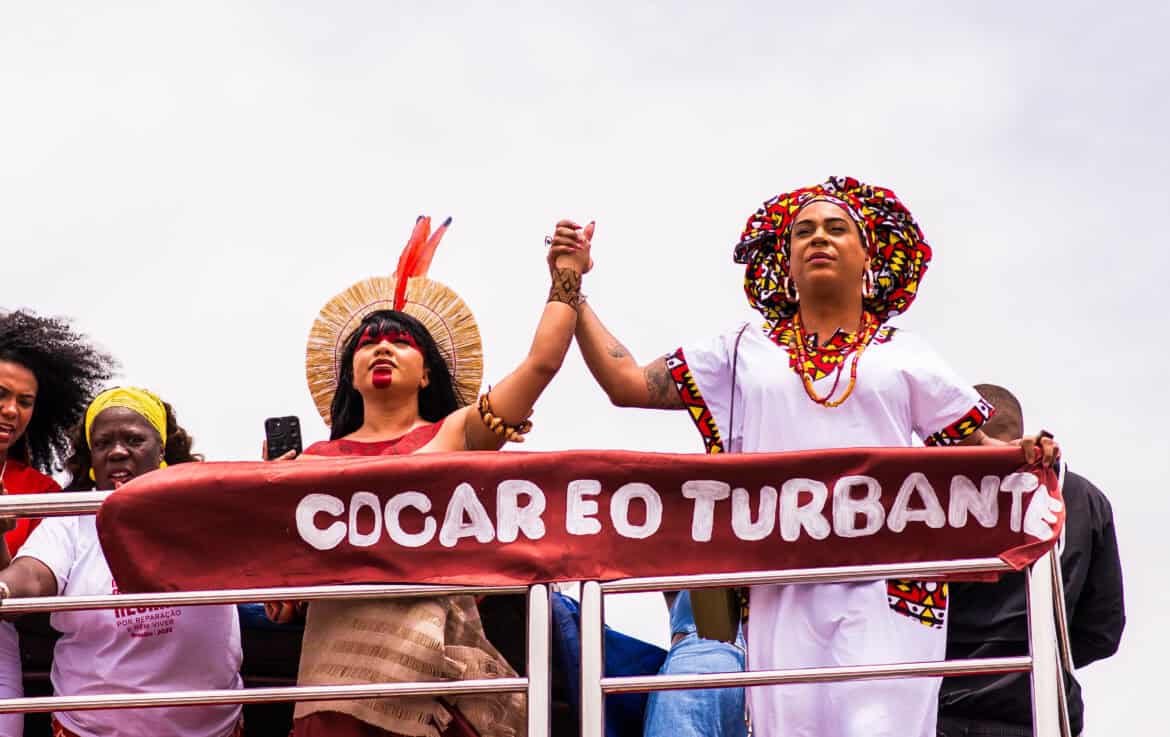 Two people holding hands in the air while looking forward in solidarity. There is a banner that says "Cocar eo turbante"