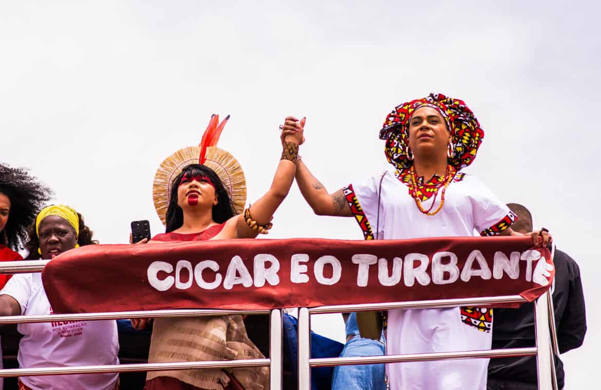 Two people holding hands in the air while looking forward in solidarity. There is a banner that says "Cocar eo turbante"