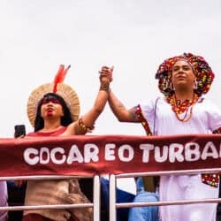 Two people holding hands in the air while looking forward in solidarity. There is a banner that says "Cocar eo turbante"