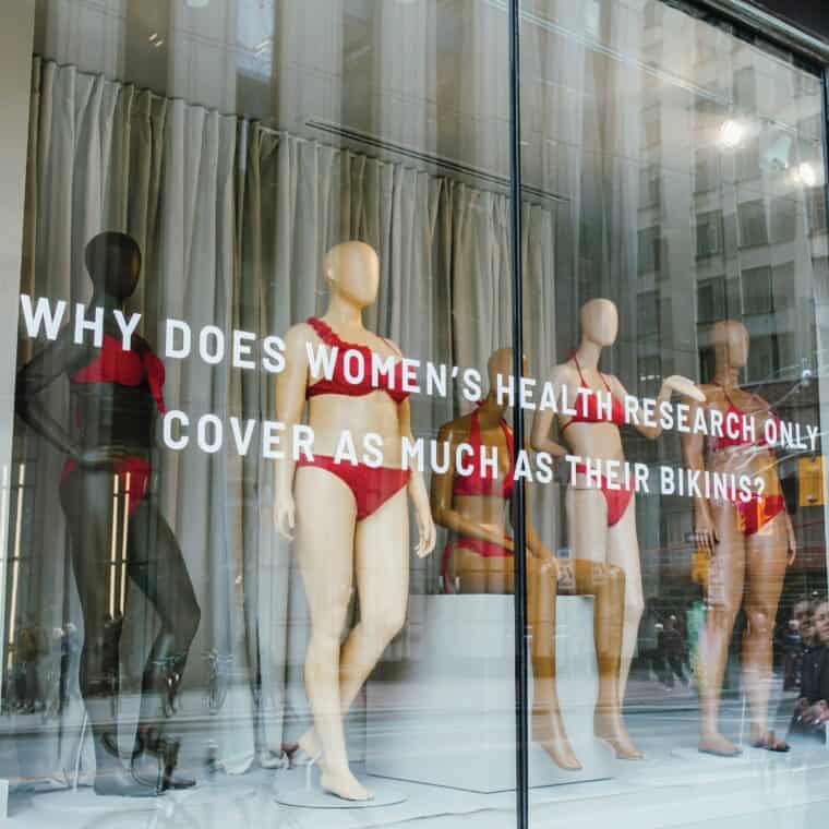 A photo shows bikini-clad mannequins displayed behind glass with the words "Why does women's health research only cover as much as their bikinis?"