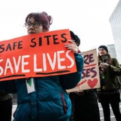 A photo of people at the court challenge rally. The person in front holds a sign that says "Safe sites save lives."