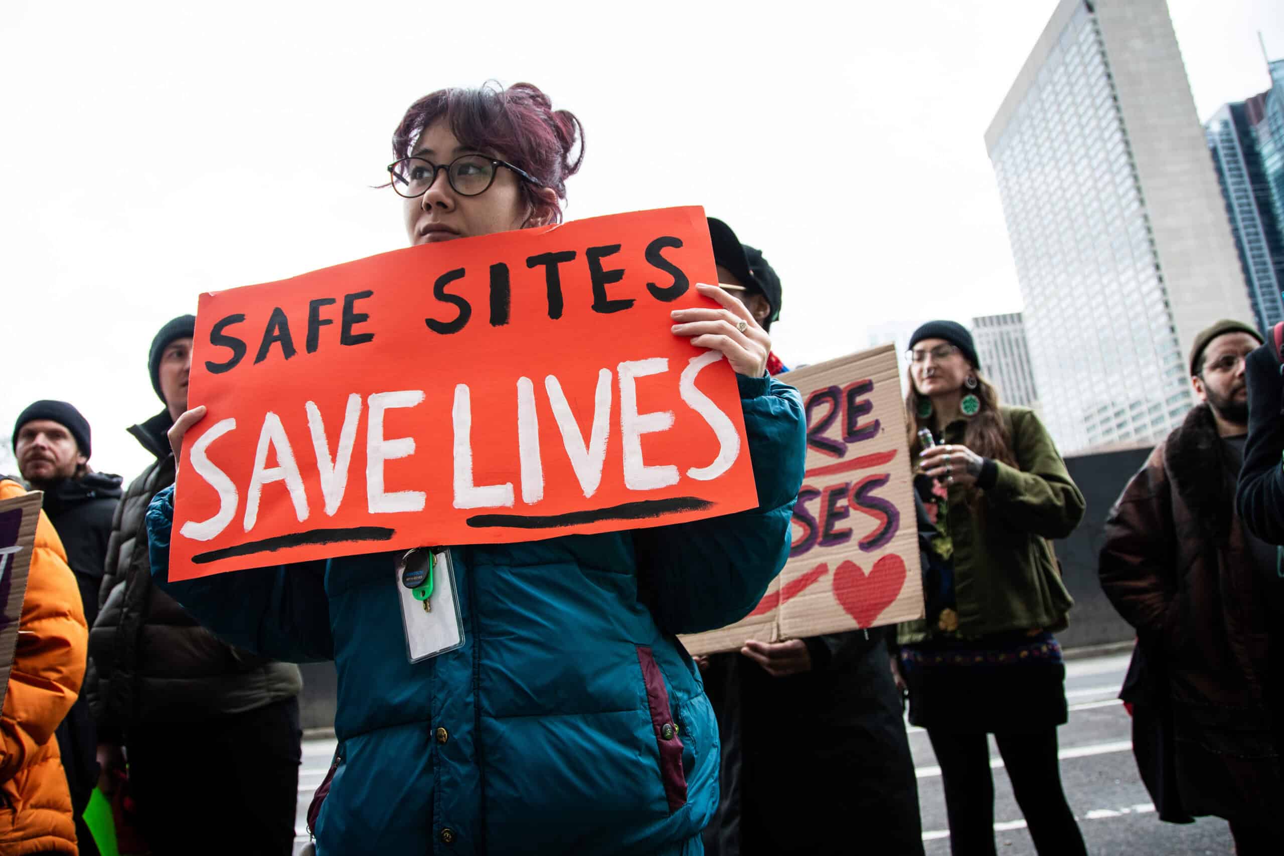 A photo of people at the court challenge rally. The person in front holds a sign that says "Safe sites save lives."